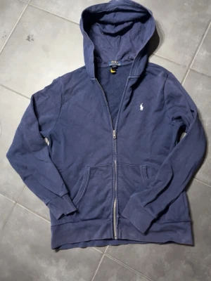 Marinblå hoodie från Polo Ralph Lauren - Snygg marinblå hoodie med dragkedja från Polo Ralph Lauren. Klassisk design med huva, kängurufickor och den ikoniska vita polospelaren broderad på bröstet. Tillverkad i mjukt bomullsmaterial som känns skönt mot huden. Perfekt för en avslappnad och stilren look.