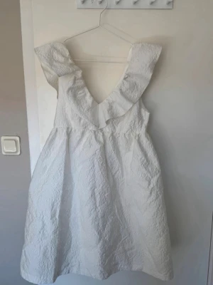 Vit kortklänning från bubbleroom - Säljer en söt vit kortklänning med djup v-ringning och bred volangkant både fram och bak. Klänningen har en lätt strukturerad yta och är ärmlös, perfekt för sommarens alla tillfällen 🥂💗