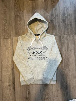 Grå hoodie från Polo Ralph Lauren - Snygg grå hoodie från Polo Ralph Lauren med stor mörkblå logga och text på bröstet🤗 använd ett par gånger men inga fel på den🙌🏻 inget äkthetsbevis tyvärr. Hör av er vid snabbaffär samt funderingar🤩
