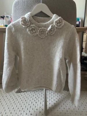 Beige stickad tröja med blommor - Supersöt beige stickad tröja med virkade blommor runt halsringningen. Tröjan har rund hals, långa ärmar och ribbade muddar vid ärmslut och nederkant. Perfekt för dig som gillar unika detaljer och en mysig stil.