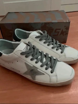 Golden Goose vita sneakers med stjärna - Golden Goose sneakers i vitt läder med grå mockastjärna på sidan och grå skosnören. Låg modell med murk grå  detalj på hälen och klassisk siluett. Märkets logga syns på plösen och sidan. Snygg streetstil som passar till det mesta. de är storlek 46 perfekta nu i vår och sommar.                                                                                     ifall du är intresserad så kan vi diskutera pris de är helt nya jag har använt dem en gång