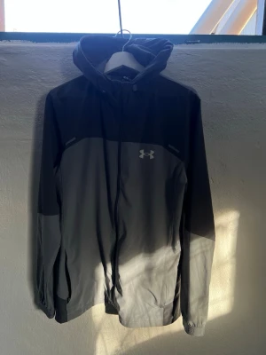 Grå vindjacka från Under Armour - Snygg grå vindjacka från Under Armour med dragkedja och huva. Jackan har två nyanser av grått, reflekterande detaljer och logga på bröstet. Perfekt för blåsiga dagar och sportiga outfits. Lätt material som andas och ger bra rörelsefrihet.