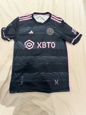 Inter Miami Messi matchtröja 23/24 - Snygg svart Inter Miami matchtröja från Adidas med rosa detaljer.  Tröjan har klubbmärke, sponsorlogga och Messis namn och nummer 10 på ryggen. Tillverkad i lätt och ventilerande material, perfekt för fotbollsträning eller support. Inte använd många gånger och inga skador