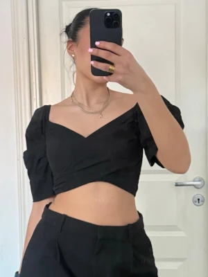 Svart croppad topp med puffärm - Trendig svart croppad topp med puffärmar och omlott-detalj framtill. Djup v-ringning och kort passform som ger en snygg siluett. Perfekt att styla med höga byxor eller kjol för en cool look.