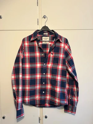 Rutig skjorta från Abercrombie & Fitch - Säljer en klassisk rutig skjorta från Abercrombie & Fitch i blått, rött och vitt. Skjortan har lång ärm, knappar framtill och en bröstficka med broderad logga. Perfekt för dig som gillar en avslappnad och cool stil.