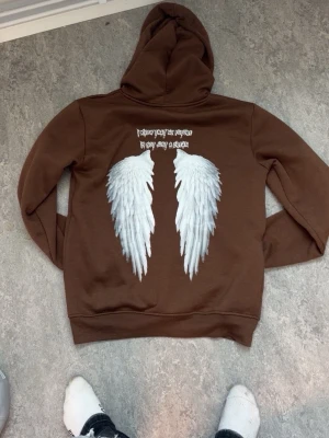 Brun hoodie med vita vingar från SHEIN - Cool brun hoodie från SHEIN med stor huva och fleecefoder. På ryggen finns ett tryck med vita änglavingar och text ovanför. Perfekt för dig som gillar unika detaljer och streetstyle. Materialet är mjukt och bekvämt, med ribbade muddar och klassisk passform.