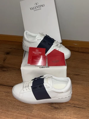 Valentino Garavani vita sneakers - Snygga vita sneakers från Valentino Garavani med en bred mörkblå panel över snörningen. Skorna har klassisk låg profil, vita snören och subtila nitar på hälen. Tillverkade i läder och har en stilren design som sticker ut.