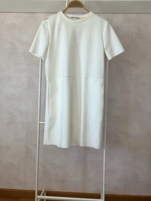 Vit kortärmad klänning från Zara  - Minimalistisk vit klänning från Zara med korta ärmar och rund hals. Klänningen har en rak passform och är sydd i ett mjukt, något tjockare material som ger en stilren look. Perfekt för dig som gillar enkel och clean stil.