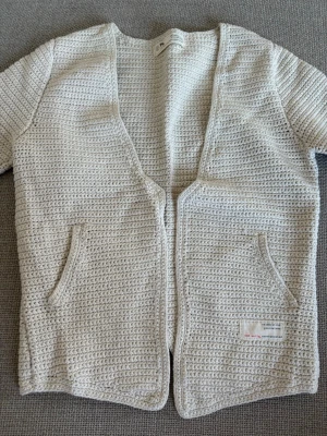 Beige virkad kofta från COS - Stilren beige virkad kofta från COS med öppen front och två fickor framtill. Koftan har korta ärmar och är gjord i ett tjockare, strukturerat material som ger en modern look. Perfekt att slänga över en t-shirt för en avslappnad vibe.