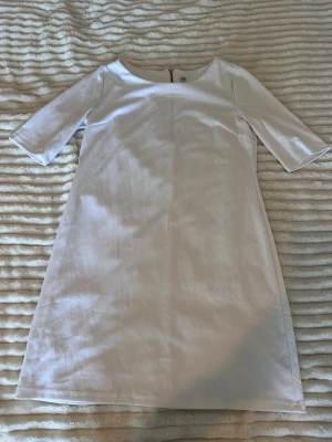 Vit enkel kortklänning med dragkedja - Stilren vit kortklänning med trekvartsärm och rund halsringning. Klänningen har en rak passform och dragkedja i ryggen för enkel påklädning. Perfekt för dig som gillar minimalistisk och clean stil.