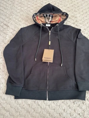 Burberry kofta - Säljer en svart hoodie från Burberry med dragkedja och klassiskt rutigt foder i huvan. Hoodien har snörning i huvan, ribbade muddar och två stora fickor framtill. Perfekt för dig som gillar stilrena och exklusiva plagg.