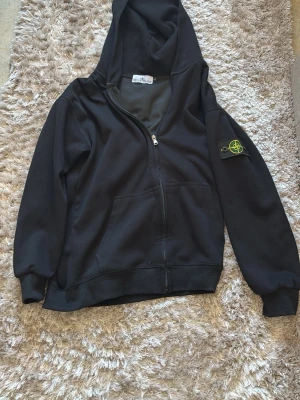 Svart hoodie från Stone Island - Svart hoodie från Stone Island med dragkedja framtill och klassisk logopatch på vänster arm. Den sitter mellan m och L dock är den större än M mindre än L