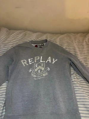 Grå sweatshirt från Replay - Snygg grå sweatshirt från Replay med tryck på bröstet där det står 'REPLAY SOUTHPORT' och ett sköldmotiv. Tröjan har rund halsringning och mjukt material på insidan. Perfekt för en avslappnad och stilren look.