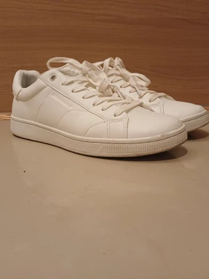 Helvita sneakers i läderlook - Stilrena helvita sneakers med lågt skaft och klassisk design. Skorna har vita snören, diskreta sömmar och ovandel i syntetläder. Perfekta för en clean och fräsch stil till jeans eller kjol.