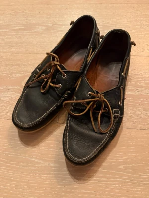 Ralph Lauren läderseglarskor med snörning - Klassiska seglarskor i läder från Ralph Lauren med bruna snören och kontrastsömmar. Skorna har rundad tå, dekorativa snören runt hälen och en låg sula. Perfekta för en avslappnad och stilren look.