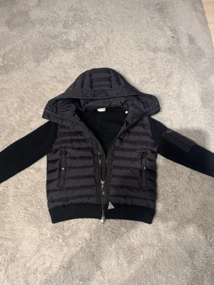 Moncler Cardigan - Säljer en svart dunjacka från Moncler med stickade ärmar och huva. Jackan har två fickor med dragkedja framtill och en extra ficka på ärmen. Perfekt för kalla vinterdagar och har en modern, sportig look.