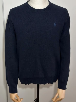 Mörkblå stickad tröja Polo Ralph Lauren - Mörkblå stickad tröja från Polo Ralph Lauren med rund halsringning och diskret blå broderad logga på bröstet. Tröjan har långa ärmar och ribbade muddar vid ärmslut och nederkant. Klassisk och enkel design i mjukt material.