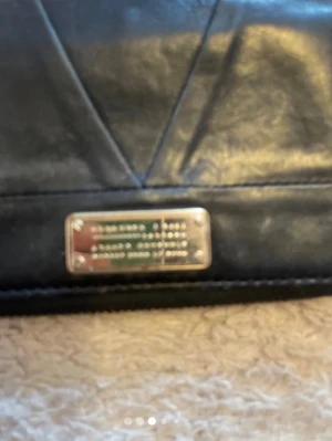 Svart plånbok från Marc Jacobs - Snygg svart plånbok från Marc Jacobs i äkta läder med silverfärgad metallbricka framtill. Insidan är vinröd och har flera kortfack, fönster för ID och en dragkedjeficka. Plånboken stängs med dragkedja runtom och har en stilren, modern design.