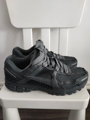 Nike Vomero sneaker - Snygga svarta sneakers från Nike med sportig design. Skorna har meshpaneler för bra ventilation, robust sula och klassisk swoosh på sidan. Färgtonen är helsvart med grå detaljer och skosnören. Perfekta för dig som gillar streetwear och sportig stil.