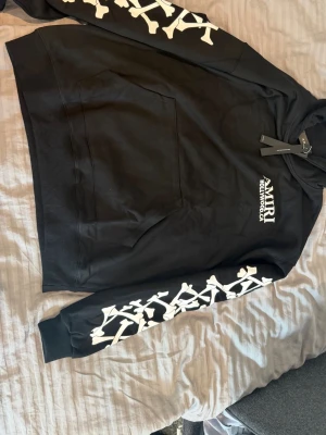 Svart Amiri hoodie med vita detaljer - Säljer en svart hoodie från Amiri med vita benmönster längs båda ärmarna och texten 'AMIRI HOLLYWOOD, CA' på bröstet. Hoodien har en klassisk huva och stor magficka framtill. Materialet är mjukt och skönt, perfekt för en streetwear-look.