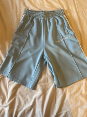 Ljusblåa/babyblåa Palm angels shorts - Jag säljer ett par babyblåa Palm angels shorts i storlek L, använt några gånger men använder inte så mycket längre. Lite skavanker precis under märket på vänstra benet med sömmen men inget större.