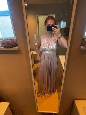 Champagnefärgad balklänning - Fin balklänning i champagnefärgad satin med omlott och djup v-ringning. Klänningen har ett plisserat fall, öppen rygg och är ärmlös med breda axelband. Klänningen är använd en gång och är i nyskick! Storlek S. Nypris: 1599