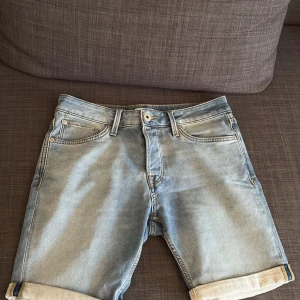 Ljusblå jeansshorts från Jack & Jones - Säljer ett par ljusblå jeansshorts från Jack & Jones med klassisk femficksdesign och uppvikta benslut. Shortsen har normal passform och är tillverkade i bomull, perfekta för varma dagar.