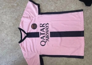 Rosa PSG matchtröja med svarta detaljer - Snygg rosa PSG matchtröja med svarta detaljer och Qatar Airways-tryck på bröstet. Tröjan har rund halsringning med svart kant, svarta ränder längs sidorna och ett PSG-märke på vänster bröst. Perfekt för fotbollsträning eller att bära till vardags.