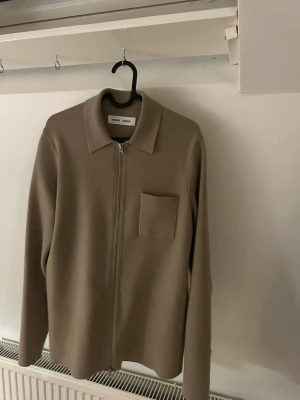 Beige overshirt från Samsoe Samsoe - Stilren beige overshirt från Samsoe Samsoe med rak passform, klassisk krage och bröstficka. Dragkedja framtill och långa ärmar. Tillverkad i ett mjukt, strukturerat material som ger en clean och modern look.