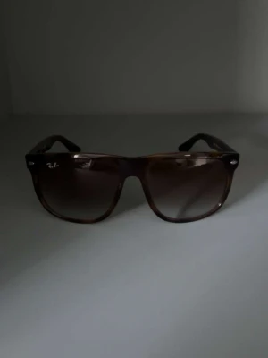 Bruna Ray-Ban solglasögon - Snygga solglasögon från Ray-Ban med klassisk fyrkantig form och bruna bågar. Linsen är mörktonad och bågarna har en subtil sköldpaddsmönstrad look. Perfekt för dig som vill ha en tidlös och cool stil. Ray-Ban-loggan finns på både lins och skalmar.