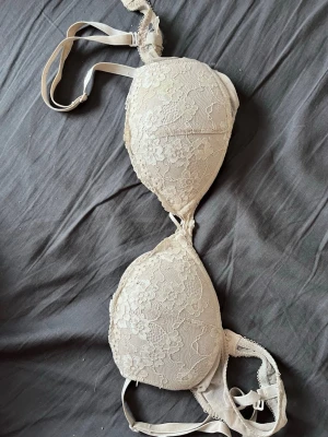 Beige spets-bh med vaddering - Snygg beige bh med vadderade kupor och vacker spetsdetalj över hela framsidan. Justerbara axelband och klassisk knäppning bak. Perfekt för dig som gillar en feminin och stilren look. Det finns ett litet hål vid banden på ryggen men det märks knappt.