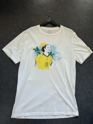 Vit t-shirt från Nakata med Ronaldinho på - Säljer en vit t-shirt med snyggt tryck av Ronaldinho på bröstet. T-shirten är från Nakata fc och har rund hals. Storlek M. Perfekt för alla fotbollsälskare. 