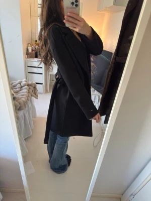 Svart klassisk kappa med knytskärp - Stilren svart kappa med knytskärp i midjan och klassisk krage. Lång modell som går ner över låren.