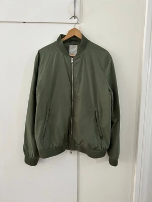 Olivgrön bomberjacka med dragkedja - Storleken är Xl, men DEN PASSAR M!!!!! Stilren olivgrön bomberjacka med klassisk passform, ribbade muddar och krage. Jackan har två snedställda fickor framtill och stängs med silverfärgad dragkedja. Perfekt för dig som gillar en enkel och clean look.