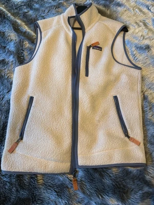 Patagonia väst - Säljer en beige fleeceväst från Patagonia i storlek M. Västen har ljusblå detaljer, tre fickor med dragkedja och snygga orange/bruna zip-pullers. 