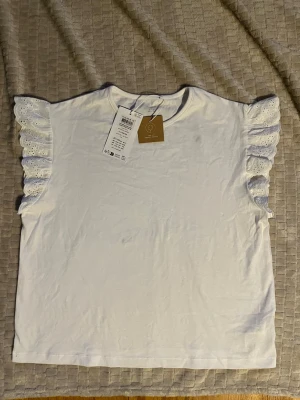 Vit topp med volangärmar och spets - Supersöt vit topp med breda volangärmar i broderad spets. Tröjan är helt ny och testat den på en gång. Det är en liten fläck vid bröstet men den går förmodligen bort om man tvättar den. Toppen har rak passform och är tillverkad i mjuk bomull. Nypris 150kr.