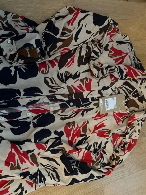 Mönstrad skjorta med knappar m - Snygg skjorta med abstrakt mönster i beige, rött, brunt och svart. Skjortan har knappar framtill och lång ärm, samt en cool spetsdetalj längs knappslån. Perfekt för dig som gillar unika plagg med färg.