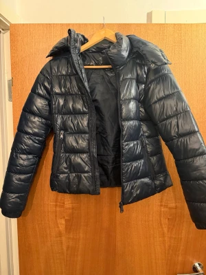 Svart pufferjacka med huva - Snygg svart pufferjacka med huva och dragkedja framtill. Jackan har quiltad design och två sidofickor med dragkedja. Perfekt för kalla dagar och ger en modern look. Materialet är glansigt och ger jackan en cool vibe.