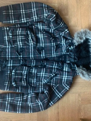 Rutig duffelkappa med fuskpäls - Snygg rutig duffelkappa i svart, vitt och rött med klassiska träknappar och huva med fuskpäls. Kappan har fickor framtill och är fodrad med mjuk teddy på insidan av huvan. Perfekt för kalla dagar och ger en cool vintagekänsla.