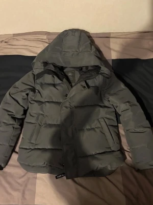 Svart dunjacka med huva från Canada goose - Snygg svart dunjacka från Canada goose Gear med stor huva och quiltad design. Jackan har päls som kommer med, två framfickor, dragkedja och tryckknappar samt en patch med logga på ärmen. Perfekt för kalla vinterdagar och riktigt varm.