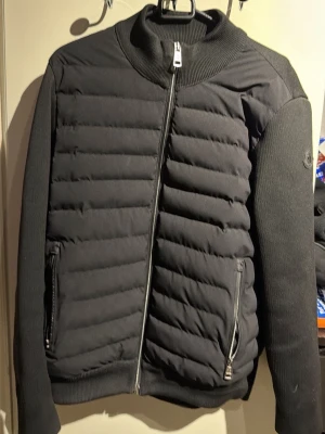 Svart pufferjacka från Moncler - Snygg svart pufferjacka från Moncler med quiltad front och stickade ärmar. Jackan har två dragkedjeförsedda fickor och ribbad krage. Moncler-logga på ärmen ger en exklusiv touch. Perfekt för kalla dagar och riktigt trendig look.