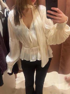 Glansig cremefärgad blus med scarf - Säljer en glansig cremefärgad blus med v-ringning och tillhörande scarf runt halsen. Blusen har långärmade puffärmar med breda muddar och ett markerat midjeparti som ger en snygg siluett.🩷 aldrig använd och har prislappen kvar, köpte för 399💕💕