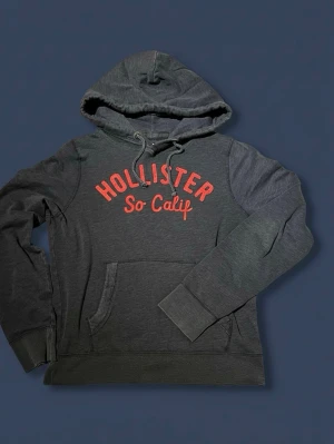 Hollister Hoodie - Mörkgrå hoodie från Hollister med en riktigt fräsh röd hollister text på bröstet. Den är mörkgrå med inga defekter förutom lite nopper som går bort lätt. Storleken är XL men passar mer som M-L eftersom den är vintage och då har krympt. Mått, längd 63 cm och bredd 48 cm
