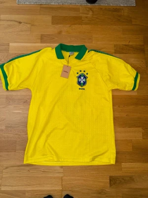 Brasilien Ronaldo 9 fotbollströja - Gul Brasilien fotbollströja från Nike med gröna detaljer, klassisk krage och tryck med Ronaldo och nummer 9 på ryggen. Broderat landslagsemblem på bröstet och gröna ränder längs ärmarna. Perfekt för fotbollsfans och samlare.