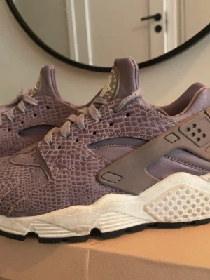 Lila Nike Air Huarache sneakers - Nike Air Huarache sneakers i lila med coolt reptilmönster på ovandelen. Skorna har en chunky vit sula och svarta detaljer undertill. Klassisk Huarache-plastdetalj runt hälen och snörning framtill. Perfekt för dig som vill sticka ut med unika sneakers.