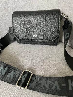 Svart axelväska från NA-KD - Snygg svart axelväska från NA-KD i fejkläder med grov struktur. Väskan har ett brett axelband med NA-KD-logga, magnetlås och innerfack med dragkedja. Perfekt för att bära det viktigaste och ger en cool streetstyle-vibe.