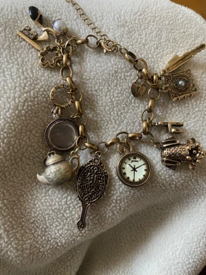Vintageinspirerat berlockarmband guld - Charmigt armband i guldfärgad metall med flera unika berlocker som tekanna, spegel, printed klocka, nyckel och bok. Kedjan är grov och armbandet har justerbar längd. Perfekt accessoar för dig som gillar detaljer och en lekfull stil.