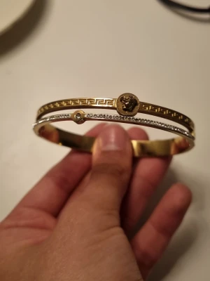 Guld- och silverfärgat armband med lejon - Snyggt armband i guld- och silverfärg från Gemstone Brilliance. Armbandet har ett lejonhuvud i mitten, grekiskt mönster och en rad vita stenar, samt en vit safir. Perfekt för dig som gillar detaljer och vill sticka ut.