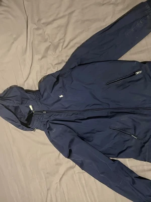 Marinblå vindjacka från Polo Ralph Lauren - Snygg marinblå vindjacka från Polo Ralph Lauren med huva och dragkedja framtill. Jackan har två snedställda fickor med dragkedja och den klassiska Polo-loggan broderad på bröstet. Tillverkad i lätt och slitstarkt material, perfekt för blåsiga dagar.