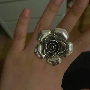 Stor silverfärgad blomring - Säljer en statementring i form av en stor blomma i silverfärgad metall. Ringen har en tydlig rosdesign med spiral i mitten och markerade kronblad. Perfekt för dig som vill sticka ut med accessoarerna och gillar unika smycken.